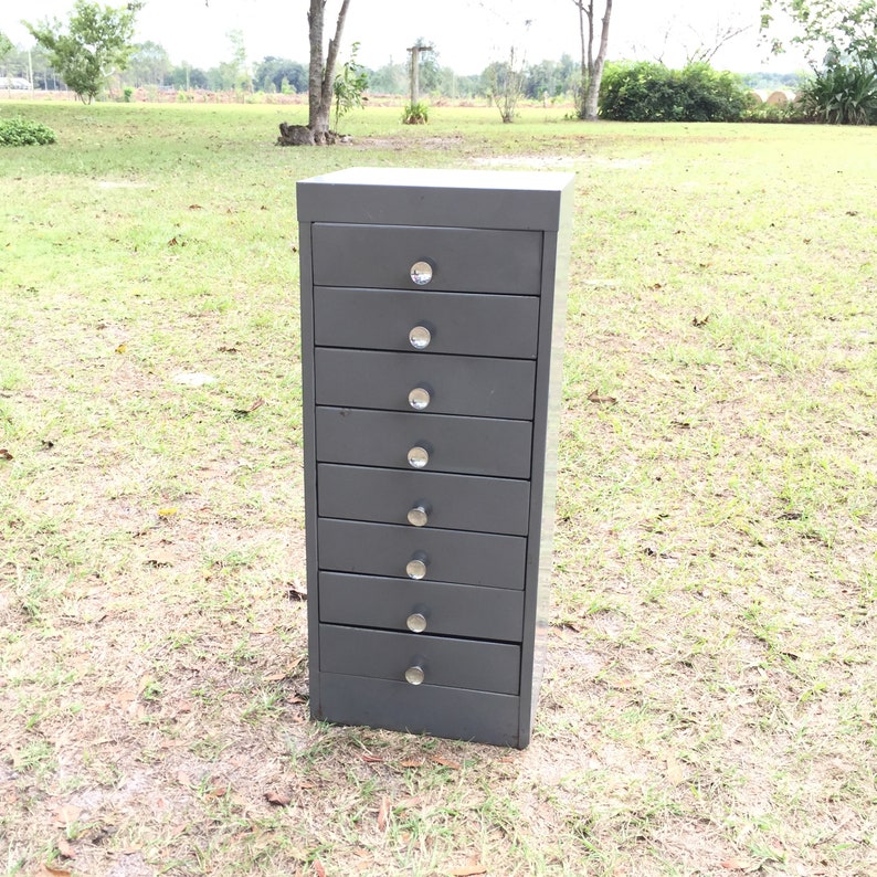 May include: A tall, gray metal cabinet with eight drawers and silver knobs. The cabinet is standing on a grassy lawn.