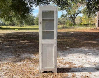 May include: A white wooden cabinet with glass doors and shelves. The cabinet has a single door on the bottom and is standing on a dirt path with brown leaves.