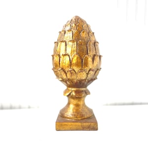 May include: A gold-colored decorative finial shaped like a pinecone with a textured, crackled finish. The object features a pointed, layered top section resting on a pedestal base with a square bottom, displaying a uniform metallic gold appearance.