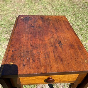 May include: A vintage wooden side table with a warm, brown finish. The table features a single drawer with a round wooden knob and turned legs. The tabletop shows signs of wear, adding to its antique charm. The table is a classic piece of furniture.
