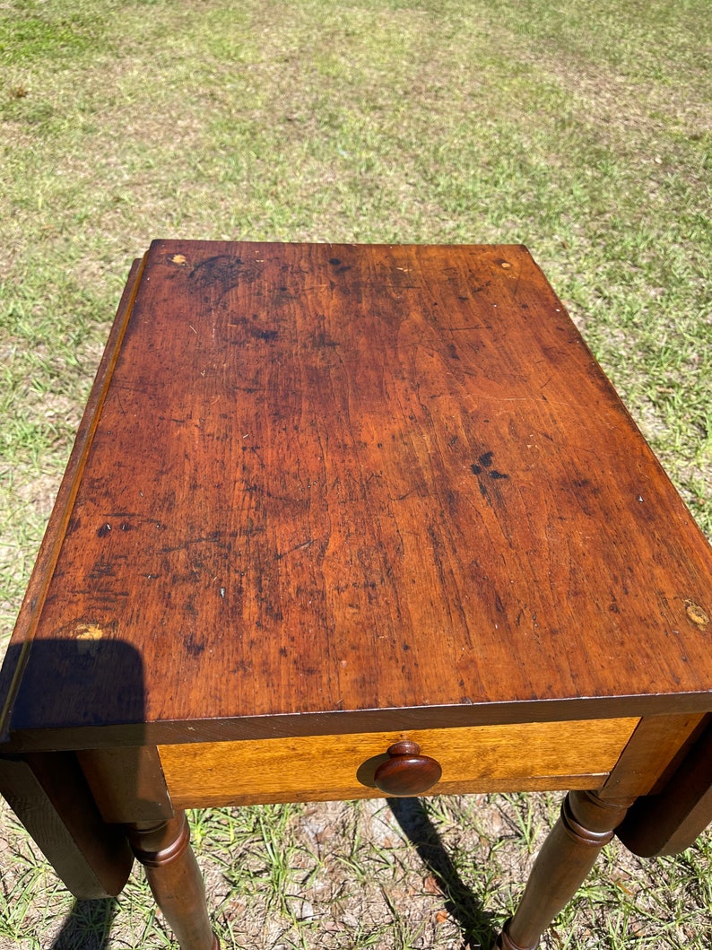 May include: A vintage wooden side table with a warm, brown finish. The table features a single drawer with a round wooden knob and turned legs. The tabletop shows signs of wear, adding to its antique charm. The table is a classic piece of furniture.