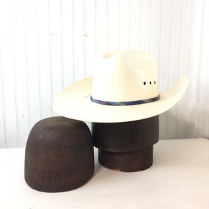 May include: A white cowboy hat with a blue and white plaid band sits on a wooden hat form. The hat form is on a white surface.