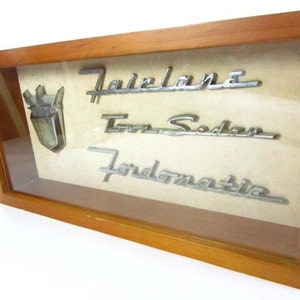 May include: A framed display of vintage car emblems, including a Ford crest, "Fairlane", "Town Sedan", and "Fordomatic".