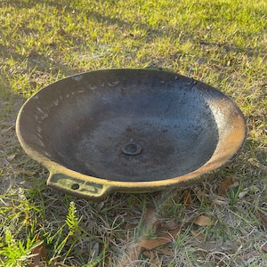 May include: A large, round, cast iron pan with a handle. The pan is dark gray and has a rough, textured surface. It is sitting on a bed of green grass.