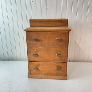 May include: A small wooden dresser with three drawers and brass oval pulls. The dresser is made of light brown wood and has a simple design.
