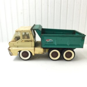 May include: A vintage green and cream toy dump truck with the word "STRETCTO" on the side. The truck has four wheels with white tires and brown rims.