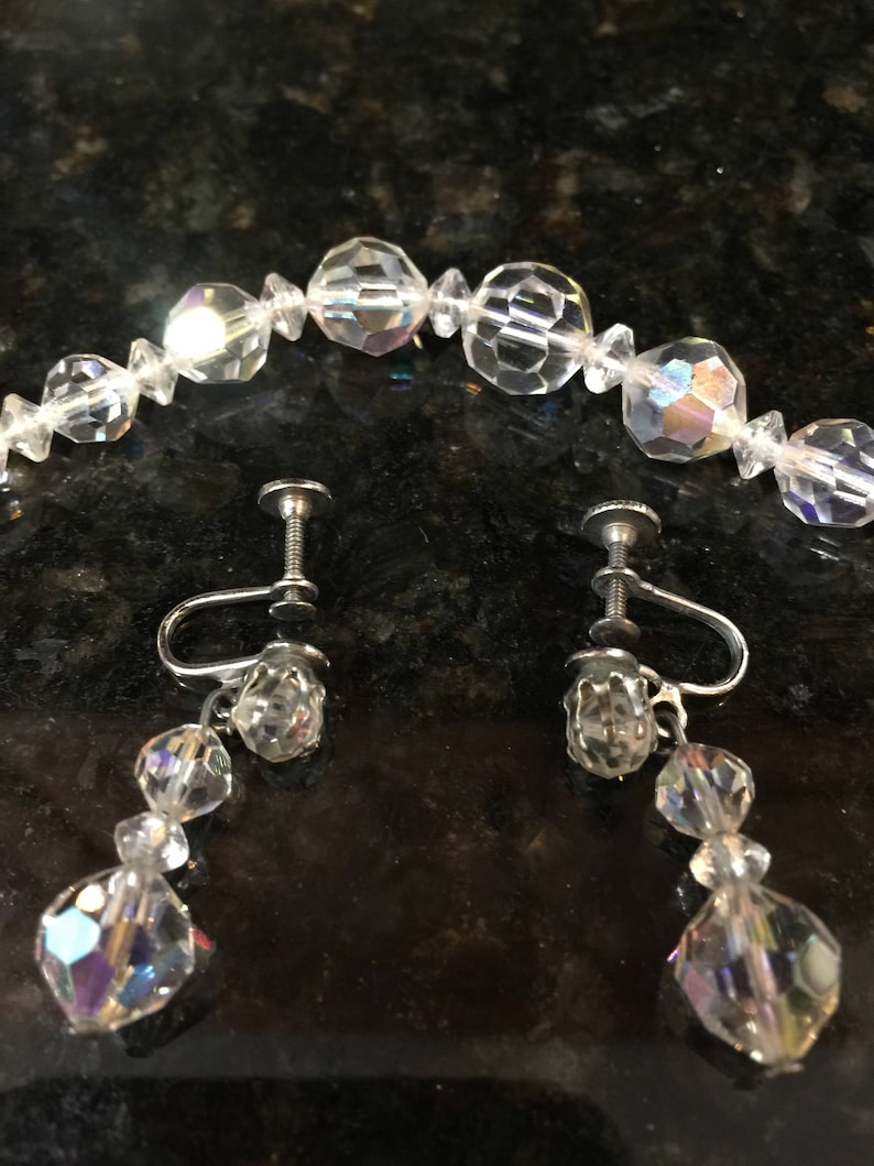 May include: A set of iridescent crystal earrings and a matching necklace. The earrings are clip-on style and feature a teardrop design with three crystal beads. The necklace is a strand of round crystal beads.