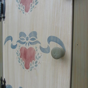 May include: A white wooden cabinet door with a blue and pink stenciled design of hearts and ribbons. The door has a round, gray knob.