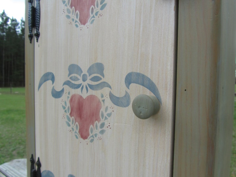 May include: A white wooden cabinet door with a blue and pink stenciled design of hearts and ribbons. The door has a round, gray knob.