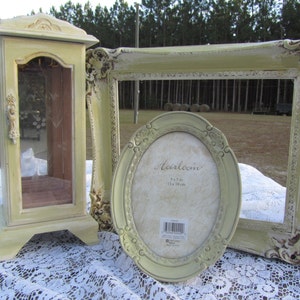 May include: Three decorative frames in a distressed white finish. The largest frame is a rectangular picture frame with ornate detailing. The middle frame is an oval picture frame with the word "Heirloom" printed on the front. The smallest frame is a tall, narrow, rectangular display case with a glass front.