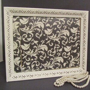 Puede incluir: Marco de imagen blanco adornado con un patr&oacute;n floral blanco y negro en el interior. Un collar de perlas blancas est&aacute; en primer plano.