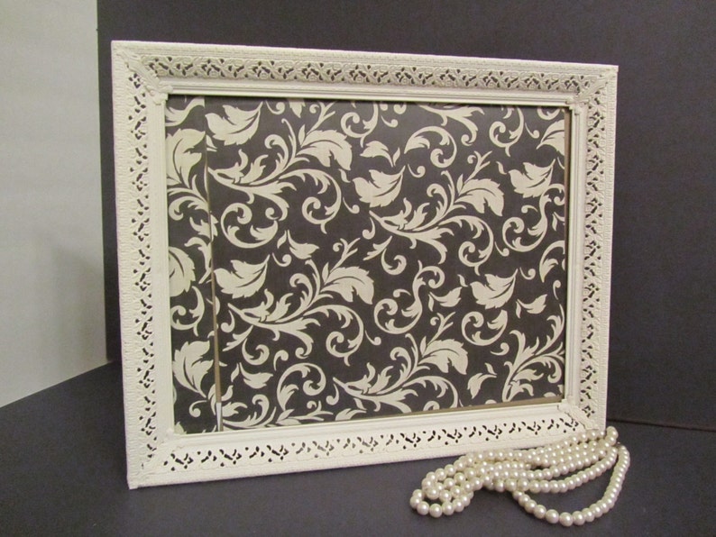 Puede incluir: Marco de imagen blanco adornado con un patr&oacute;n floral blanco y negro en el interior. Un collar de perlas blancas est&aacute; en primer plano.