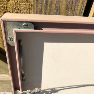 Puede incluir: Primer plano de la parte posterior de un marco de imagen rosa claro con un alambre de metal para colgar sujeto al marco. El marco tiene una pieza de esquina de metal plateado con un tornillo.