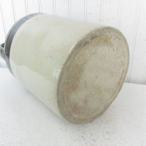 May include: A large, white ceramic crock with a dark blue rim. The crock has a textured surface and is missing a handle.