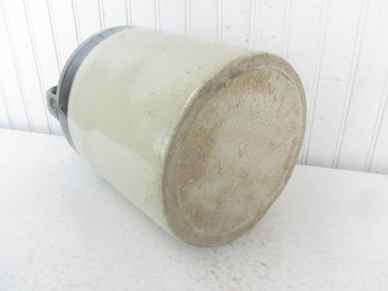May include: A large, white ceramic crock with a dark blue rim. The crock has a textured surface and is missing a handle.
