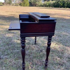 May include: A dark brown wooden writing desk with a drop-down writing surface and multiple drawers. The desk has turned legs and is sitting on a grassy lawn.