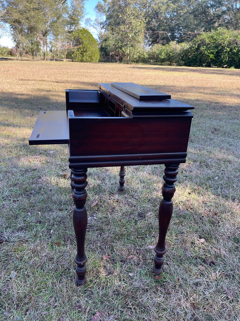 May include: A dark brown wooden writing desk with a drop-down writing surface and multiple drawers. The desk has turned legs and is sitting on a grassy lawn.