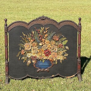 May include: A decorative fireplace screen with a black background and a large floral arrangement. The flowers are in shades of yellow, red, and orange, with a blue vase. The screen has a dark brown frame and stands on two legs.
