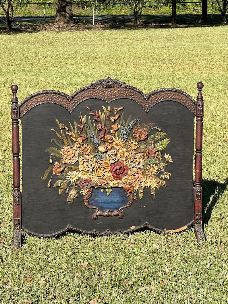 May include: A decorative fireplace screen with a black background and a large floral arrangement. The flowers are in shades of yellow, red, and orange, with a blue vase. The screen has a dark brown frame and stands on two legs.