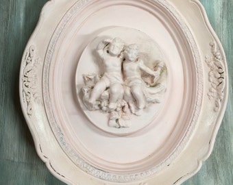 Könnte beinhalten: Ovale Wanddekoration im Vintage-Stil. Der Rahmen ist in verwittertem Weiß gehalten, mit einem rosa Innenbereich. In der Mitte befindet sich eine weiße Basrelief-Skulptur von zwei Engeln. Der Hintergrund ist in einem verwitterten Türkis gehalten.