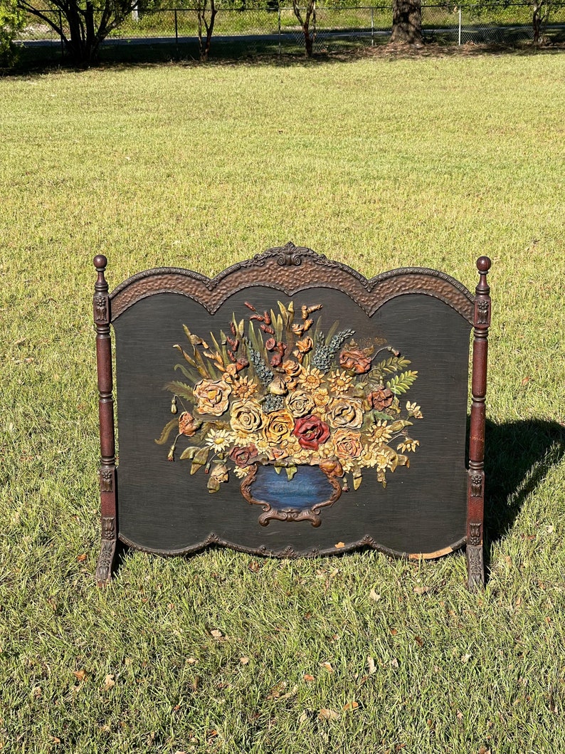 May include: An antique fireplace screen with a black background and a detailed floral arrangement in shades of yellow, red, and orange. The screen has a decorative, ornate frame and dark brown wooden legs.