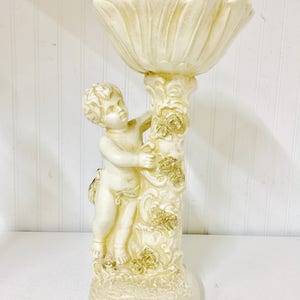 May include: Off-white cherub figurine holding a decorative, petal-shaped bowl. The cherub is sculpted with detailed features and is attached to a column adorned with floral accents. The base is round and textured, creating a vintage aesthetic.