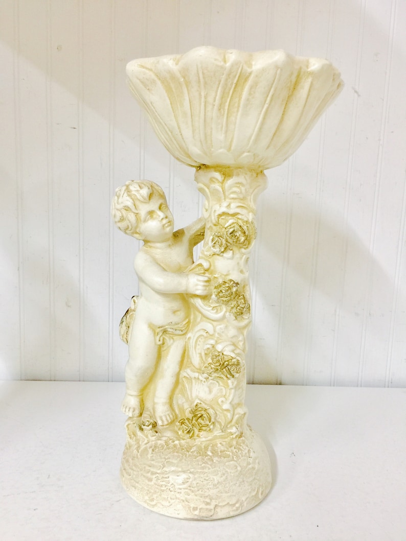 May include: Off-white cherub figurine holding a decorative, petal-shaped bowl. The cherub is sculpted with detailed features and is attached to a column adorned with floral accents. The base is round and textured, creating a vintage aesthetic.