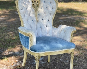 May include: A white and blue upholstered armchair with a carved wooden frame. The chair has a tufted back and seat, and the fabric is a soft velvet. The chair is in a garden setting.