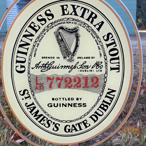 K&ouml;nnte beinhalten: Ein Vintage-Schild f&uuml;r Guinness Extra Stout mit einem goldenen und wei&szlig;en runden Design. Das Schild zeigt eine Harfe, den Text "Guinness Extra Stout" und die Worte "Brewed in Ireland by Arthur Guinness Son & Co Ltd as their trade mark and label".
