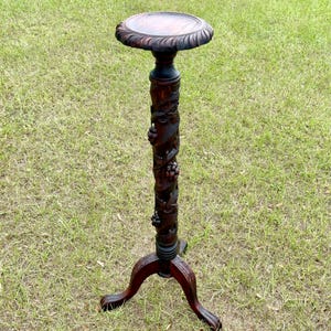 May include: A dark brown, carved wooden plant stand with a round top and three curved legs. The central column features carved grape vine details. The stand is approximately 30 inches tall.