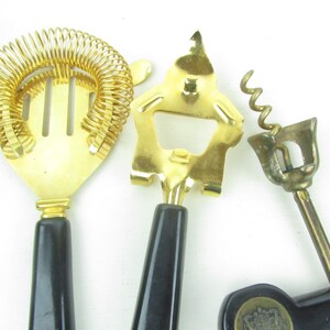 Op de afbeelding: Een set van drie gouden bargereedschappen met zwarte handvatten. De gereedschappen omvatten een cocktailscheider, een flesopener en een kurkentrekker.