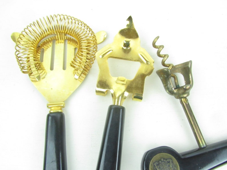 Op de afbeelding: Een set van drie gouden bargereedschappen met zwarte handvatten. De gereedschappen omvatten een cocktailscheider, een flesopener en een kurkentrekker.