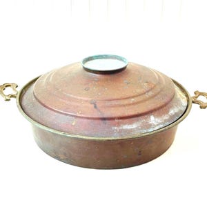 May include: A vintage copper cooking pot with a lid. The pot has a round shape with a slightly flared rim and two ornate brass handles. The lid has a central knob and concentric rings. The copper has a reddish-brown patina.