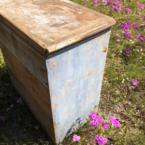 May include: A vintage wooden chest with a blue painted exterior. The chest has a hinged lid and is made of weathered wood. The chest is sitting on a bed of pink flowers.