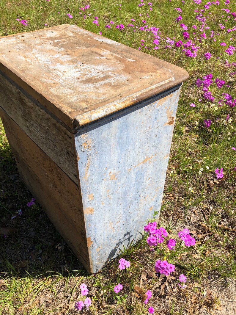 May include: A vintage wooden chest with a blue painted exterior. The chest has a hinged lid and is made of weathered wood. The chest is sitting on a bed of pink flowers.