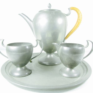 May include: A pewter tea set with a teapot, creamer, sugar bowl, and tray. The teapot has a brown wicker handle. The set is on a white background.