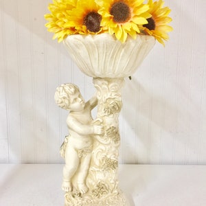 May include: A cream-colored cherub-themed vase holding bright yellow sunflowers. The vase has a detailed design with a cherub figure supporting the floral bowl. The sunflowers have brown centers and yellow petals, creating a vibrant contrast.