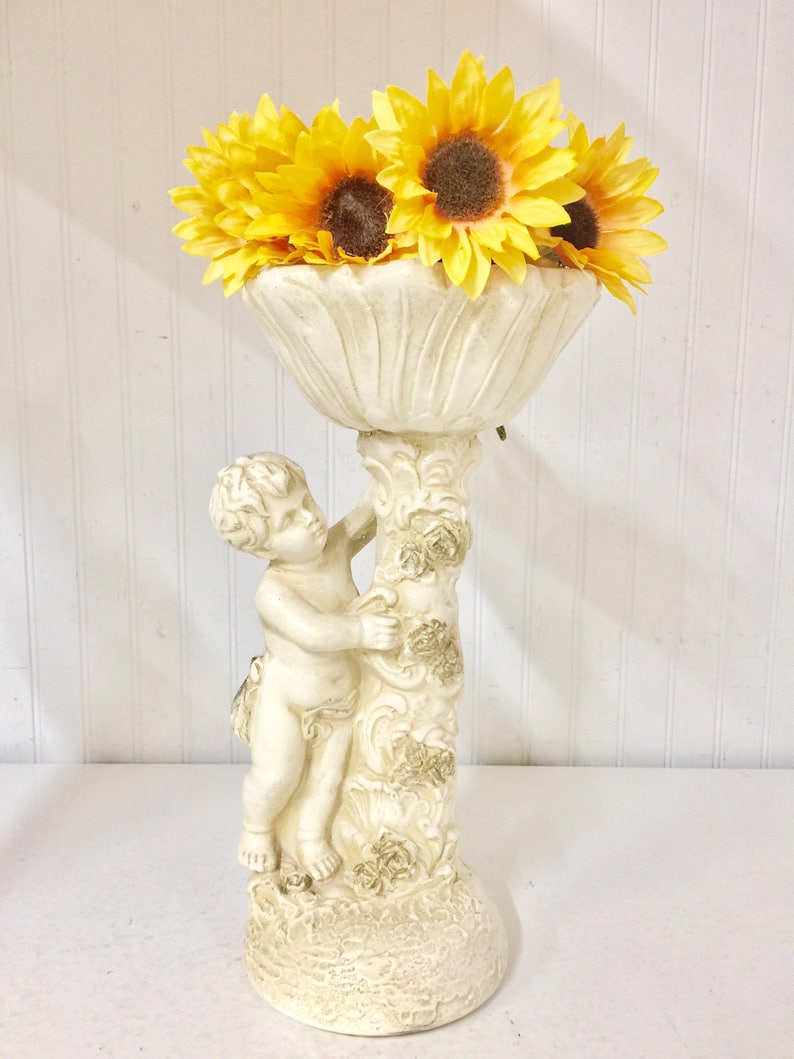May include: A cream-colored cherub-themed vase holding bright yellow sunflowers. The vase has a detailed design with a cherub figure supporting the floral bowl. The sunflowers have brown centers and yellow petals, creating a vibrant contrast.