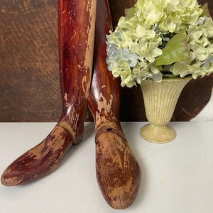 May include: A pair of vintage wooden shoe forms with a distressed red finish. The forms are shaped like a foot and have a rounded toe. They are sitting on a white surface with a vase of green flowers in the background.