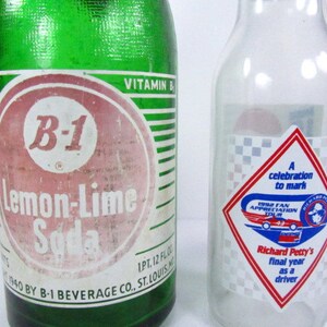 May include: A green glass bottle with a white label that reads "B-1 Lemon-Lime Soda". The bottle is next to a clear glass bottle with a red, white, and blue diamond-shaped sticker that reads "A celebration to mark 1992 Fan Appreciation Tour Richard Petty's final year as a driver".