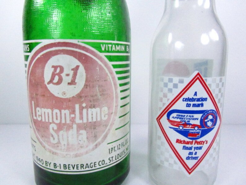May include: A green glass bottle with a white label that reads "B-1 Lemon-Lime Soda". The bottle is next to a clear glass bottle with a red, white, and blue diamond-shaped sticker that reads "A celebration to mark 1992 Fan Appreciation Tour Richard Petty's final year as a driver".