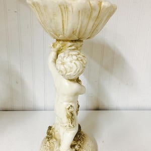 May include: A cream-colored cherub figurine holding a shell-shaped bowl. The statue is made of a light-colored material with a textured finish. The cherub has curly hair and is standing on a textured base.