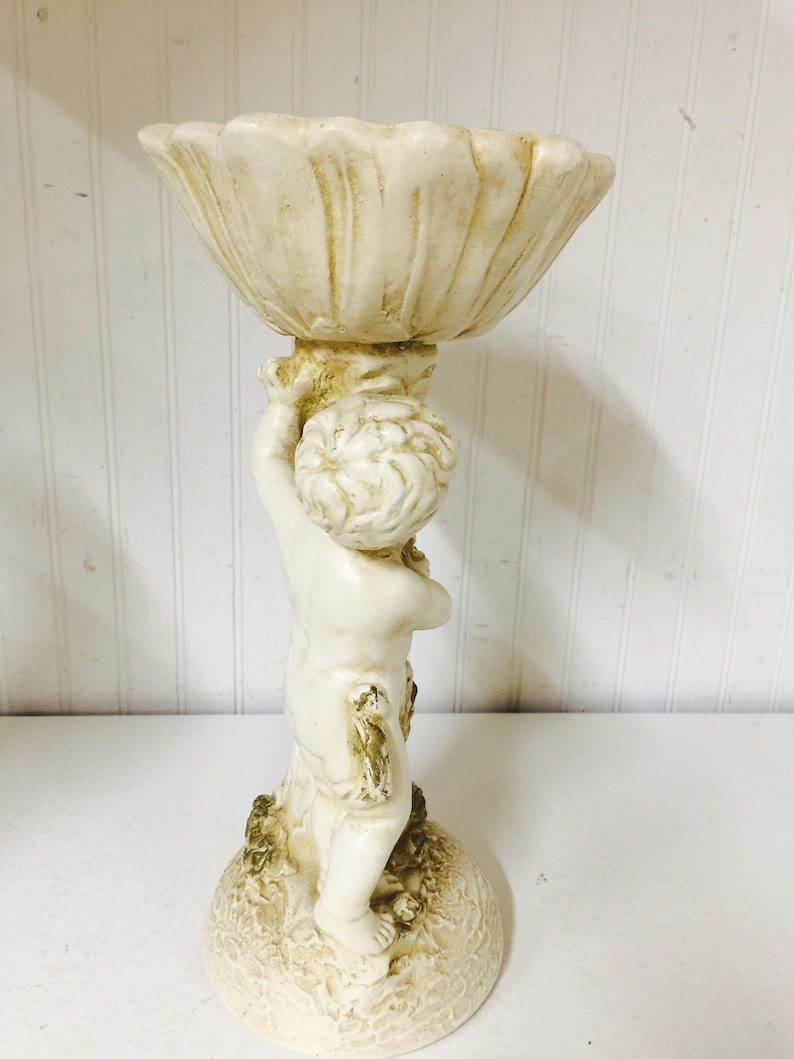 May include: A cream-colored cherub figurine holding a shell-shaped bowl. The statue is made of a light-colored material with a textured finish. The cherub has curly hair and is standing on a textured base.