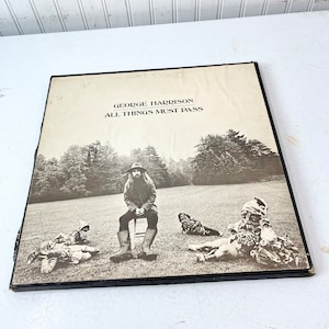 May include: A black and white photo of George Harrison on the cover of his album "All Things Must Pass". The photo shows Harrison sitting on a stool in a field with three other people lying on the ground.