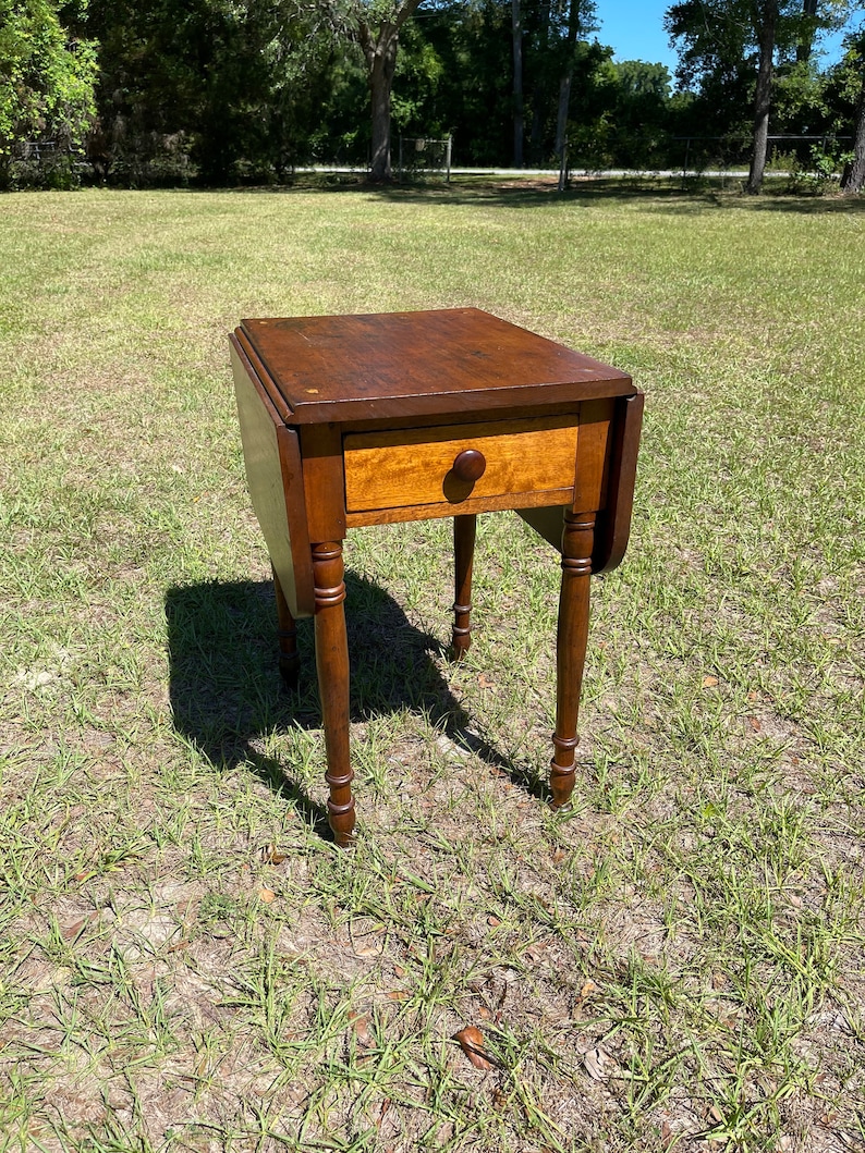May include: A small, antique wooden side table with a dark brown top and a single drawer. The drawer has a round wooden knob. The table has four slender, turned legs. The table's dimensions are approximately 24 inches high, 20 inches wide, and 20 inches deep.