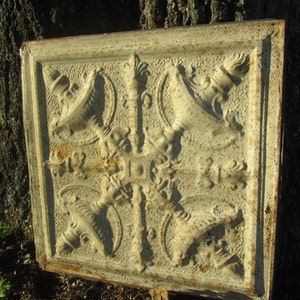 May include: A white metal ceiling tile with a decorative embossed design featuring a repeating pattern of stylized floral motifs and a central circular design.