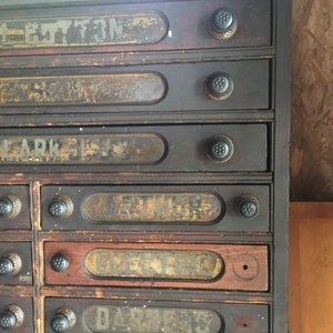 May include: Antique wooden cabinet with eight drawers, each with a label. The labels are faded and worn, but the words "BUTTON", "ARK", "ARTHUR", "ELEC", and "DARNERS" are visible. The cabinet has black metal knobs.