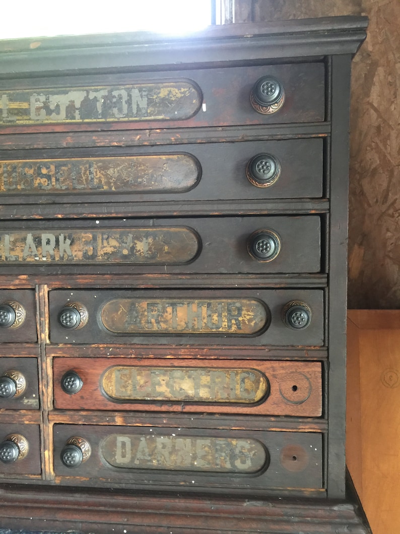 May include: Antique wooden cabinet with eight drawers, each with a label. The labels are faded and worn, but the words "BUTTON", "ARK", "ARTHUR", "ELEC", and "DARNERS" are visible. The cabinet has black metal knobs.