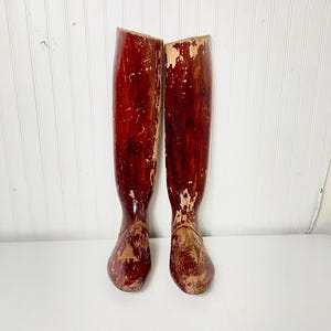 May include: A pair of vintage wooden boot forms, painted in a deep red color with some areas of the paint worn away, revealing the natural wood underneath. The forms are shaped like a boot and have a rounded toe.