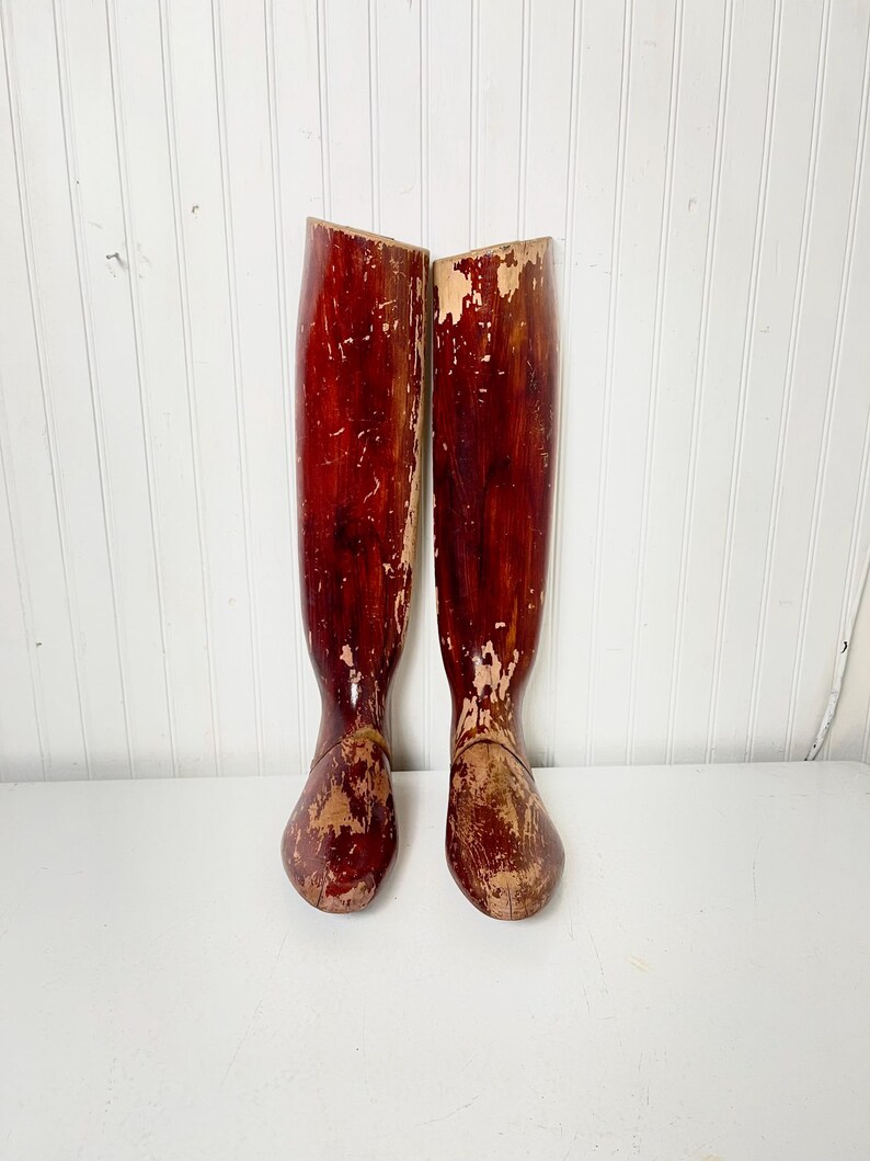 May include: A pair of vintage wooden boot forms, painted in a deep red color with some areas of the paint worn away, revealing the natural wood underneath. The forms are shaped like a boot and have a rounded toe.